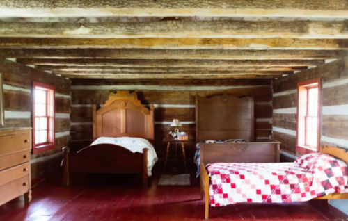 A rustic bedroom with two wooden beds, a dresser, and a small table, featuring log cabin walls and red wooden floors.