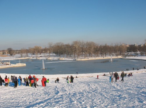 People sledding and playing in the snow near a frozen pond with fountains, surrounded by trees under a clear blue sky.