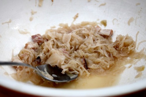 A bowl of sauerkraut with a spoon, featuring finely shredded cabbage and bits of meat in a light brine.