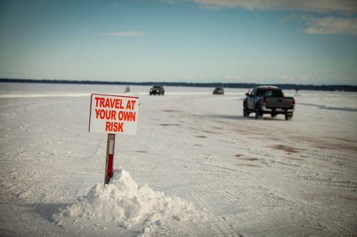 A sign reading "Travel at Your Own Risk" stands in snow, with vehicles driving on a frozen surface in the background.