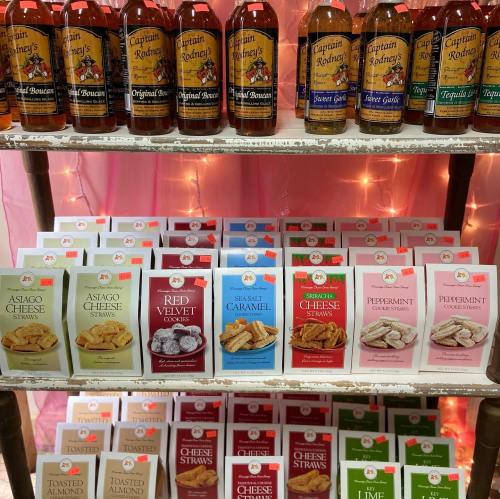 A display of various bottles and snack boxes, including cookies and cheese straws, on a shelf with a pink backdrop.