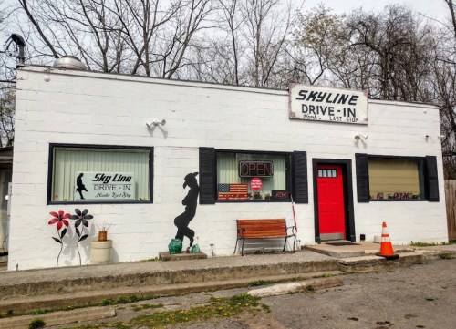 A small drive-in restaurant with a white exterior, featuring a silhouette of a woman and an "Open" sign in the window.