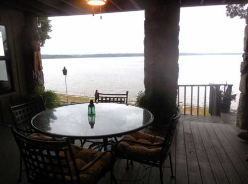 A cozy outdoor seating area with a glass table overlooking a calm lake and distant shoreline.