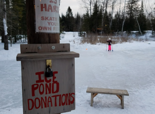 A wooden donation box labeled "Ice Pond Donations" near a frozen pond where a person skates in winter attire.