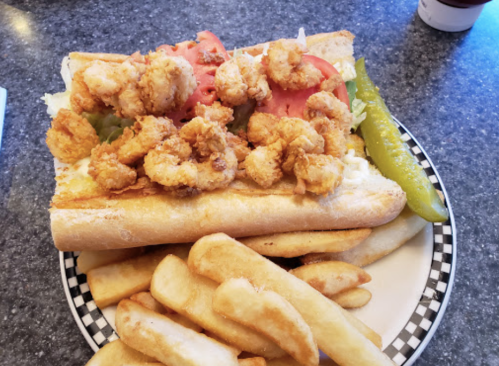 A shrimp po' boy sandwich topped with lettuce, tomatoes, and pickles, served with a side of golden fries.