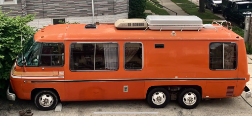 An orange vintage RV parked on a residential street, featuring large windows and a rooftop air conditioning unit.