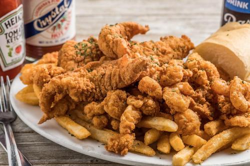 A plate of crispy fried seafood, including shrimp and fish, served with French fries and a roll, with condiments in the background.