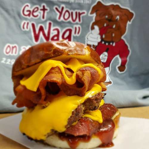 A stacked burger with cheese, bacon, and barbecue sauce on a pretzel bun, with a cartoon bulldog shirt in the background.