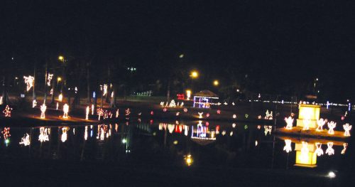 Colorful holiday lights reflect on a calm pond at night, surrounded by trees and festive decorations.