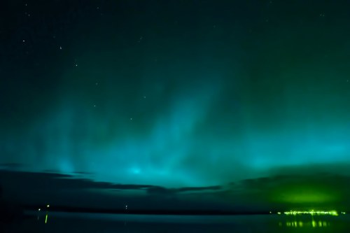 A serene night sky illuminated by vibrant green and blue auroras over a calm lake, with distant lights on the shore.