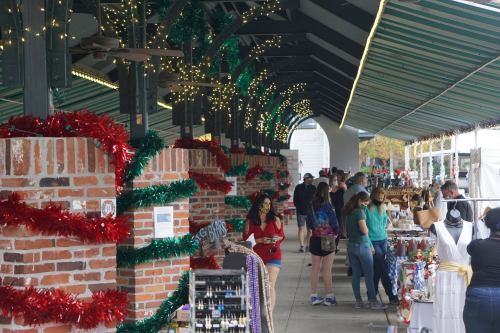 A festive market scene with people browsing stalls decorated with holiday garlands and lights under a striped canopy.