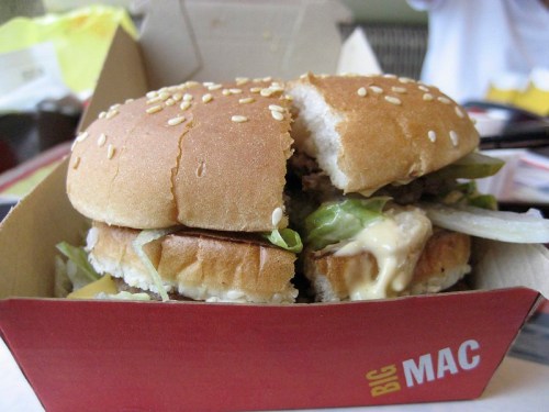 A close-up of a Big Mac burger with sesame seed bun, lettuce, cheese, and sauce, partially bitten.