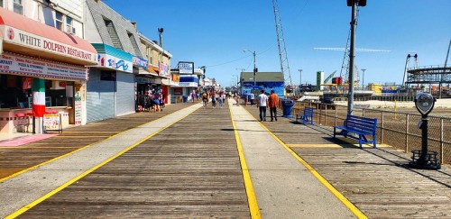 A bustling boardwalk with shops and attractions, lined with wooden planks and people enjoying a sunny day.