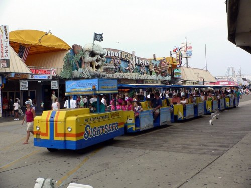 A colorful sightseeing tram on a boardwalk, surrounded by shops and people enjoying a sunny day.