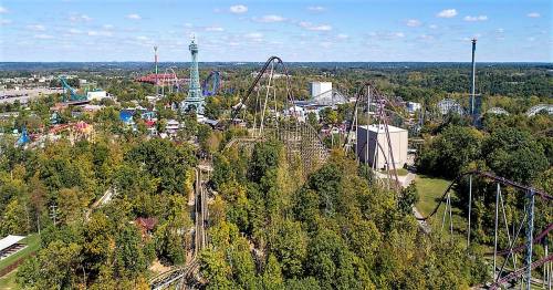 Aerial view of an amusement park featuring roller coasters, rides, and lush greenery under a blue sky.