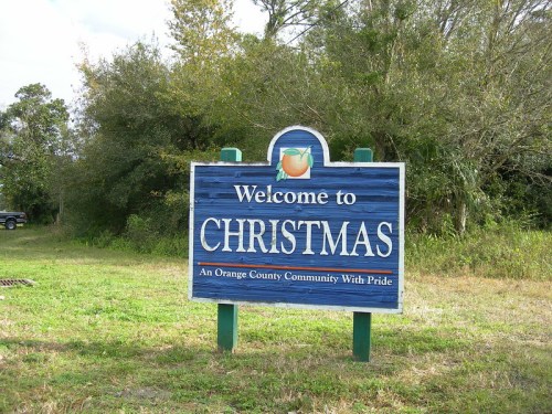 Sign reading "Welcome to Christmas" with an orange graphic, set in a grassy area surrounded by trees.