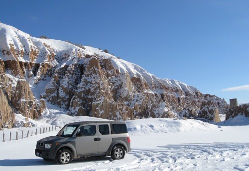 A silver SUV parked on a snowy landscape with rugged, snow-covered cliffs in the background under a clear blue sky.