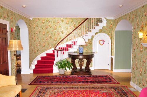A bright foyer with floral wallpaper, a red carpeted staircase, a decorative table, and a potted plant.