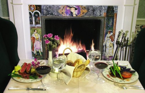 A cozy dining setup with two plates of gourmet food, wine glasses, and a roaring fireplace in the background.
