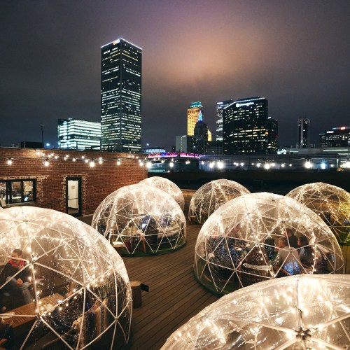 Rooftop view of illuminated igloo-like structures with city skyline at night, featuring tall buildings and string lights.