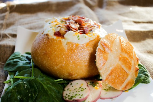 A bread bowl filled with creamy soup, topped with bacon, served on a bed of spinach and garnished with radish slices.