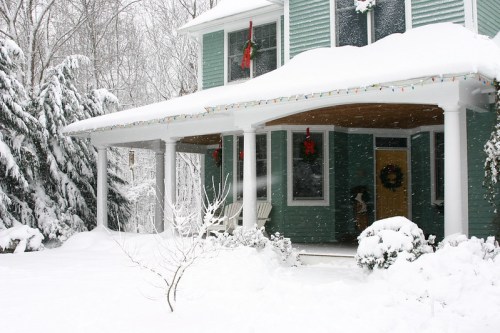 A snow-covered house with a porch, decorated with wreaths and holiday lights, surrounded by a winter landscape.