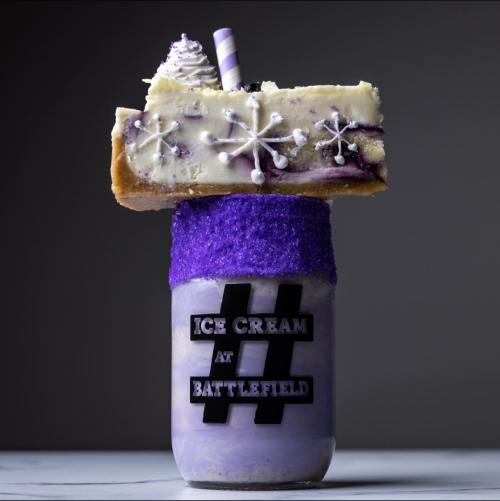 A tall dessert featuring purple ice cream topped with a slice of cheesecake, whipped cream, and a striped straw.
