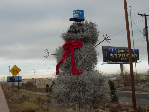 A large snowman made of tumbleweeds, wearing a blue hat and a red bow, stands beside a road with a billboard in the background.