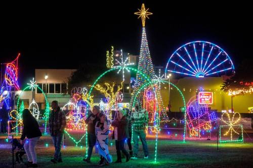 Colorful holiday lights illuminate a festive park with a large Christmas tree, a ferris wheel, and families enjoying the scene.