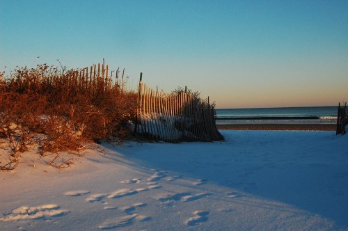 A snowy beach scene with wooden fencing and footprints leading to the calm ocean under a clear blue sky.