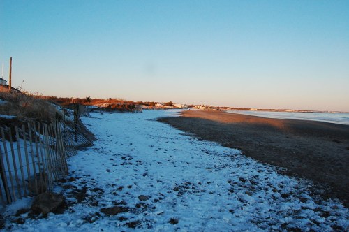 A serene beach scene with snow on the sand, a clear sky, and distant buildings along the shoreline.