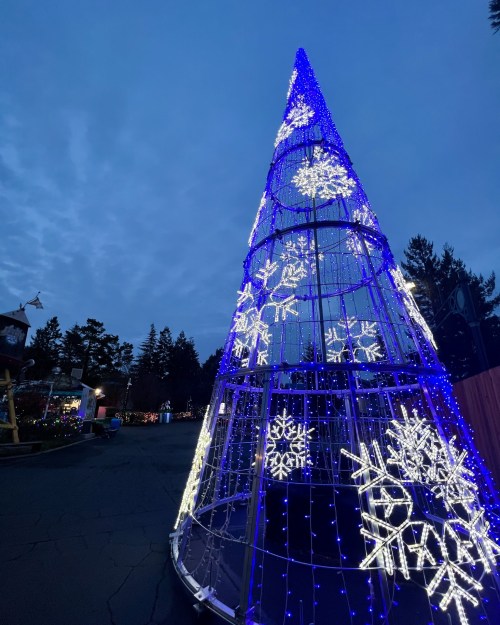 A tall, illuminated Christmas tree with blue and white lights, featuring snowflake designs, against a twilight sky.