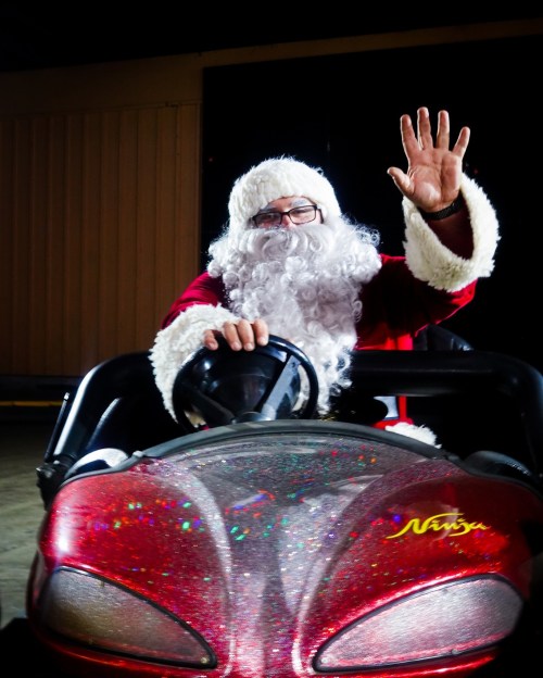 Santa Claus waves from a colorful go-kart, wearing his traditional red suit and glasses, with a cheerful expression.