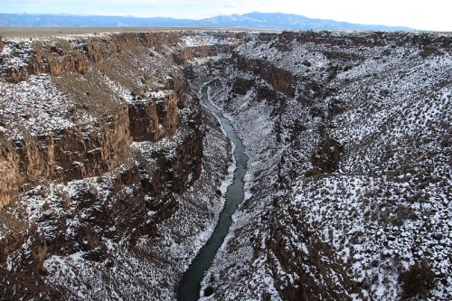A snowy canyon with steep rocky walls and a winding river at the bottom, surrounded by distant mountains.