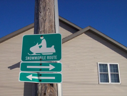 Sign indicating a snowmobile route with directional arrows, mounted on a utility pole in front of a house.