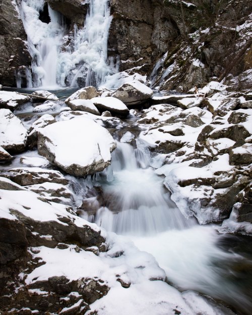 A serene winter scene featuring a waterfall cascading over rocks, surrounded by snow-covered terrain.