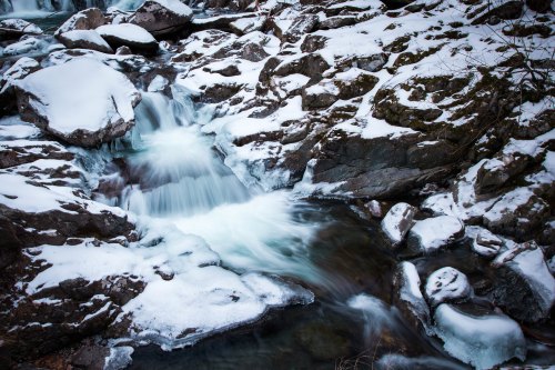 A serene winter scene featuring a flowing stream surrounded by snow-covered rocks and icy patches.