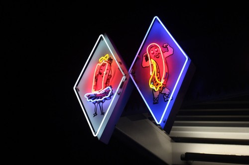 Neon sign featuring two cartoon hot dogs, one in a tutu and the other flexing muscles, against a dark background.