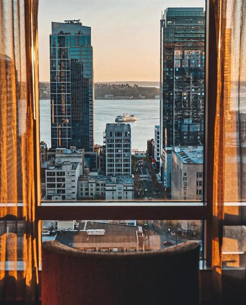 View from a hotel window showcasing city skyscrapers and a ferry on the water at sunset. Warm light filters through curtains.