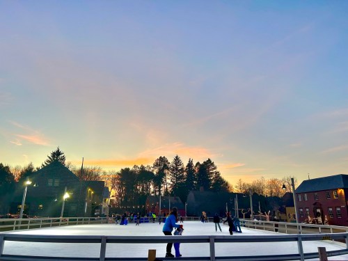 A winter scene of an outdoor ice skating rink at sunset, with skaters enjoying the ice and trees in the background.