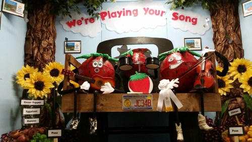 A colorful display featuring musical fruit characters, sunflowers, and a sign that says "We're Playing Your Song."