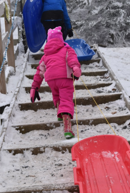 A child in a pink snowsuit pulls a red sled up snowy stairs, with another sled in the background.