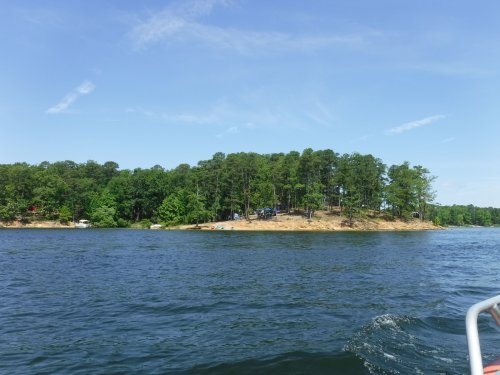 A serene lake scene with a wooded shoreline under a clear blue sky, featuring calm water and a sandy beach area.