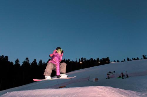 A snowboarder in a pink jacket jumps off a ramp at dusk, with trees and lights in the background.