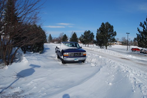 A blue truck partially buried in snow, surrounded by trees and a clear blue sky. Snow covers the ground.