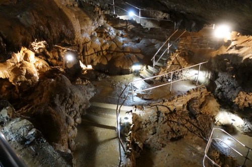A dimly lit cave interior with stone steps and metal railings, showcasing rock formations and a wet floor.