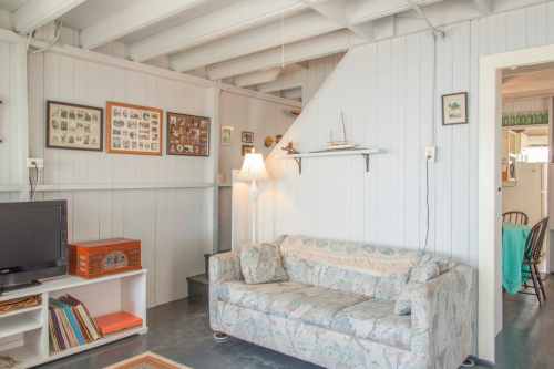 Cozy living room with a patterned couch, lamp, framed photos, and a shelf, featuring light wood paneling and a warm atmosphere.