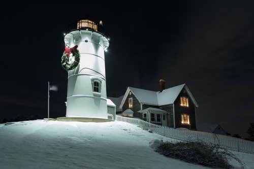 A snowy lighthouse at night, adorned with a festive wreath, next to a cozy, warmly lit house.