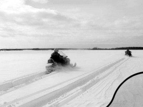Two snowmobiles ride across a snowy landscape, leaving tracks in the fresh snow under a cloudy sky.