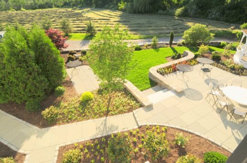 Aerial view of a landscaped garden with pathways, seating areas, and neatly arranged greenery. Fields in the background.